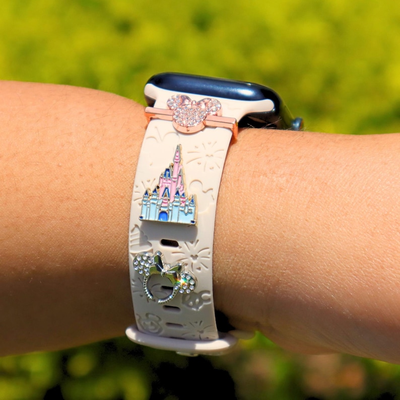 May include: A white Apple Watch band with a pink and white castle charm, a pink and white Mickey Mouse charm, and a silver Mickey Mouse charm with rhinestones.