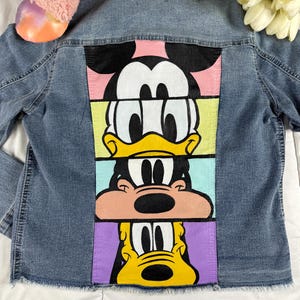 Puede incluir: Una chaqueta vaquera con un diseño pintado de colores con Mickey Mouse, Donald Duck y Goofy. Los personajes están apilados uno encima del otro, con Mickey Mouse en la parte superior y Goofy en la parte inferior.
