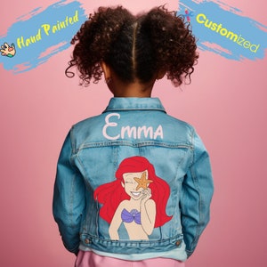 Könnte beinhalten: Hellblaue Jeansjacke mit handgemaltem Design von Arielle, der Meerjungfrau. Der Name "Emma" ist über der Grafik aufgedruckt. Die Jacke ist kundenspezifisch und hat einen klassischen Kragen und einen Knopfverschluss.