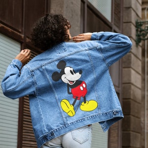 Könnte beinhalten: Hellblaue Jeansjacke mit einer großen Mickey Mouse-Grafik auf dem Rücken. Die Zeichentrickfigur ist in ihrer klassischen Pose dargestellt und trägt rote Shorts, gelbe Schuhe und weiße Handschuhe. Die Jacke hat einen klassischen Kragen und Knopfverschlüsse.