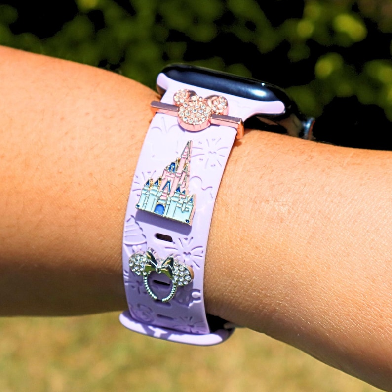 May include: A lavender silicone Apple Watch band with a gold-tone clasp and three charms: a pink Mickey Mouse head, a pink castle, and a pink bow.