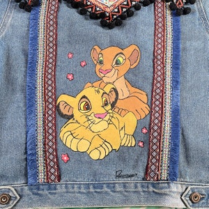 May include: A blue denim jacket with a colorful embroidered trim and a painted image of Simba and Nala from The Lion King. The image is surrounded by pink flowers.