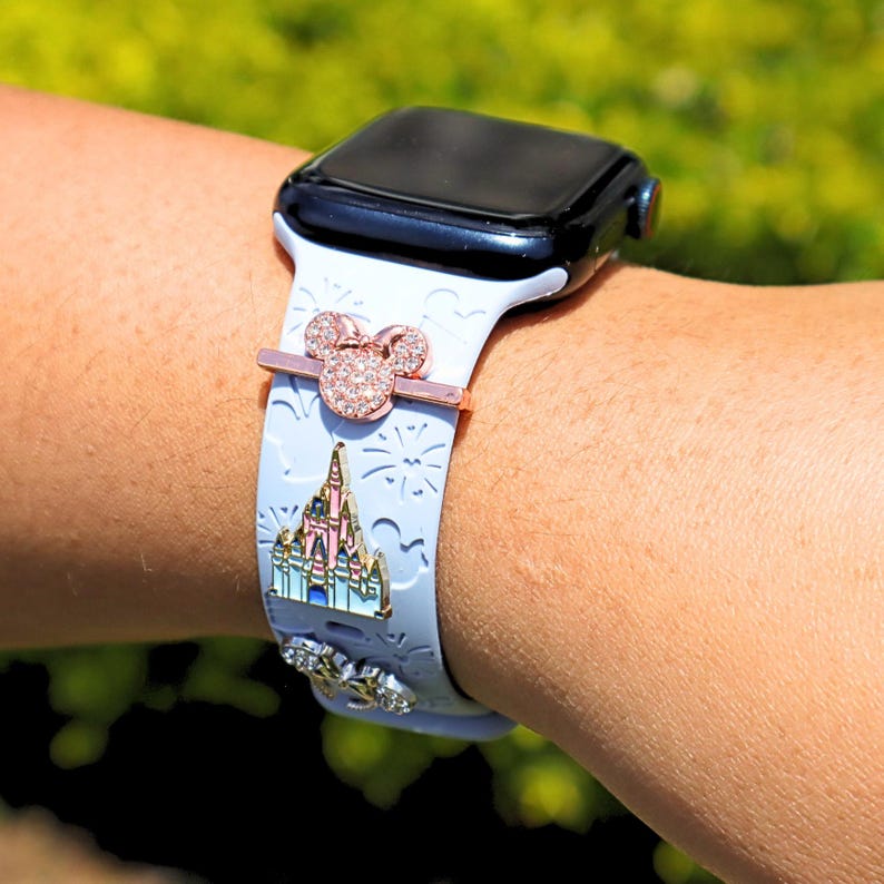 May include: A white Apple Watch band with a pink and white castle, fireworks, and a Minnie Mouse charm. The band is decorated with rhinestones.