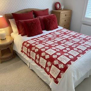 May include: A bed with a red and white quilt, red pillows, and a wicker headboard. The quilt features a geometric pattern with floral embroidery. The bed is made up with a white bed skirt and a white comforter. The room has a neutral color scheme.