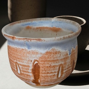 May include: A handmade ceramic bowl with a unique design. The bowl features a light blue rim, transitioning to a band of orange-brown, and a white interior. The exterior has vertical white accents. The bowl is likely used for serving food or as a decorative piece.