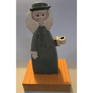 May include: A ceramic figurine of a person wearing a dark green coat and hat, holding a small object. The figurine has a light-colored face and a halo-like shape behind the head. It is mounted on a small, rectangular wooden base.