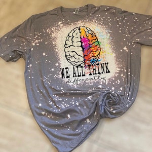 May include: Gray t-shirt with a white bleach splattered design. A colorful brain graphic is centered on the shirt with the text "WE ALL THINK differently".