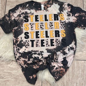 May include: A black and white tie-dye t-shirt with the word "STEELERS" in yellow and black block letters repeated four times. The shirt has a distressed, bleached look, with a knot at the bottom.