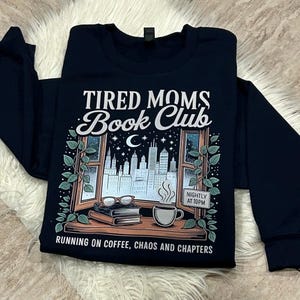 May include: Navy blue sweatshirt with the text "TIRED MOMS Book Club" above an illustration of a window with a city skyline, books, and a coffee cup. The text "RUNNING ON COFFEE, CHAOS AND CHAPTERS" is below the illustration.