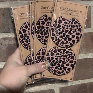 May include: Set of four brown and pink leopard print car coasters. The coasters are packaged in clear plastic and have a brown paper backing with the text "Car Coasters Handmade with love".