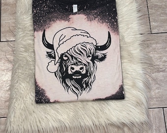 May include: A black and white graphic of a Highland cow wearing a Santa hat on a bleached light pink t-shirt.