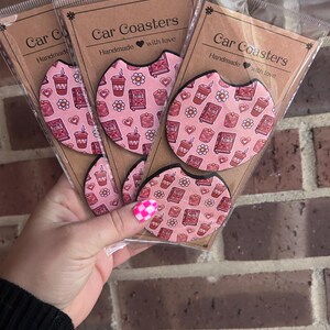 May include: Set of four pink and white car coasters with a coffee cup and book pattern. The coasters are handmade with love and feature the text "Car Coasters".