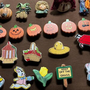 May include: A collection of decorative pins and magnets with fall and Halloween themes. Includes pumpkins, a scarecrow, a red wagon, a basket of apples, a straw hat, a sign that says "Keep Off The Grass", and Easter-themed pins.