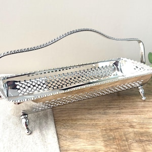 May include: A rectangular silver-toned metal basket with a lattice-patterned side design, a beaded rim, and a single arched handle. The basket is supported by four small, rounded feet and features a polished, reflective finish throughout the structure.