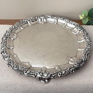 Peut inclure: Un plateau de service rond en métal argenté doté d'un bord surélevé orné de volutes complexes et de détails floraux. La surface centrale du plateau présente un motif gravé subtil et l'objet repose sur de petits pieds décoratifs.