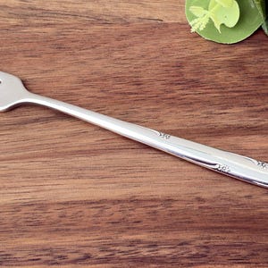 Puede incluir: Un tenedor plateado con tres púas, apoyado sobre una superficie de madera. El tenedor tiene un diseño decorativo en el mango. La imagen también incluye un paño azul claro y hojas verdes.