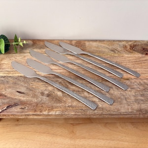 May include: Six stainless steel butter knives with textured handles are arranged diagonally on a wooden board. The knives have a simple design, with a wide, rounded blade and a textured handle. The board is made of wood.