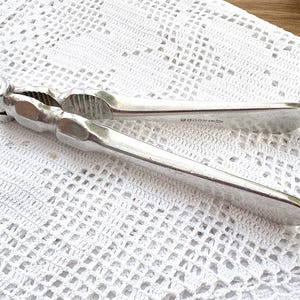 Peut inclure: Un casse-noix argenté avec un design articulé, posé sur une nappe blanche à tissage ouvert. Le casse-noix a une poignée texturée et un mécanisme de fissuration denté. Le design est simple et fonctionnel.