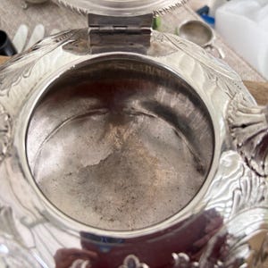 May include: A silver-colored metal vessel with an open circular top and a hinged lid. The exterior surface features intricate embossed floral and leaf patterns. The interior is smooth and metallic, showing signs of age and use with a reflective finish.