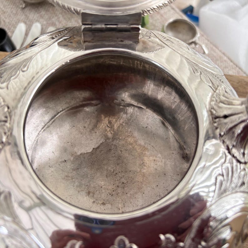 May include: A silver-colored metal vessel with an open circular top and a hinged lid. The exterior surface features intricate embossed floral and leaf patterns. The interior is smooth and metallic, showing signs of age and use with a reflective finish.