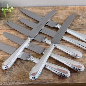 May include: A collection of silver-colored knives with a decorative handle design, arranged on a wooden surface. The knives have a shiny, reflective appearance, and the handles feature a beaded pattern. The blades are smooth and sharp.