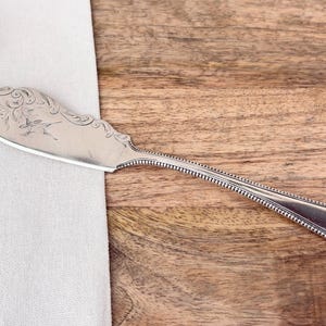 Può includere: Coltello da burro argentato con un motivo floreale decorativo sulla lama. Il manico ha un bordo perlato. Il coltello poggia su un panno bianco e una superficie in legno.