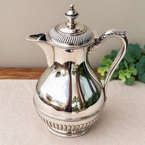 May include: A polished silver-toned metal coffee pot featuring a rounded body, a hinged lid with a finial, and a curved handle with leaf-like detailing. The base and lid are decorated with beaded trim and vertical fluting, creating a reflective surface.