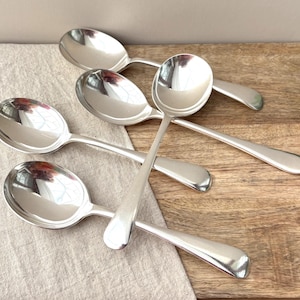 May include: A set of four polished silver-toned metal spoons with rounded bowls and smooth, plain handles. The spoons are arranged in an overlapping pattern, showcasing their reflective surfaces and simple, functional design for dining use.