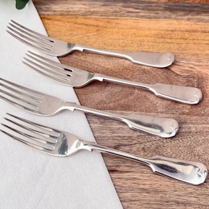 May include: Four silver forks arranged diagonally on a wooden surface, with a white cloth in the lower left corner. The forks have a classic design with tines and a rounded handle. The wood has a warm, brown tone.