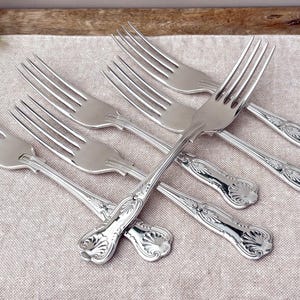 Könnte beinhalten: Ein Set aus fünf silberfarbenen Metallgabeln, die fächerförmig angeordnet sind. Jede Gabel hat eine polierte Oberfläche mit einem dekorativen Muschelmotiv am Griffende und einen Kopf mit vier Zinken. Die Metalloberflächen reflektieren das Licht.