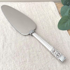 May include: A silver-toned cake server with a smooth, triangular blade and a cylindrical handle. The handle features vertical ridges and a detailed, embossed floral pattern near the base, providing a textured grip for this serving utensil.
