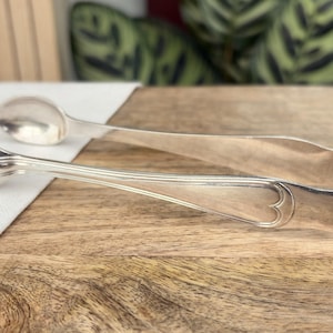 Può includere: Pinze per zucchero argentate con estremità a forma di cucchiaio e un design decorativo a forma di cuore. Le pinze sono appoggiate su una superficie di legno con un panno bianco e un fogliame verde sullo sfondo. Un classico utensile da portata.