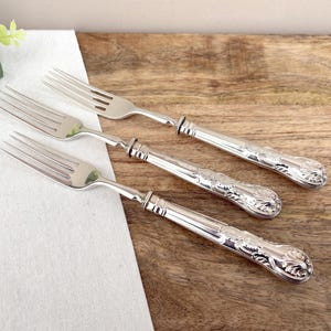 Op de afbeelding: Drie zilveren vorken met sierlijke bloemmotieven op de handvatten liggen op een houten oppervlak. De vorken hebben tanden en zijn gemaakt van een glanzend, reflecterend metaal. De handvatten hebben een gedetailleerd, reliëfpatroon.