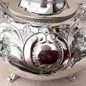 May include: A polished silver teapot featuring intricate embossed floral and scrollwork patterns across its rounded body. The center displays a circular cartouche engraved with the cursive letter A. It stands on small, ornate feet with a rope-like border.