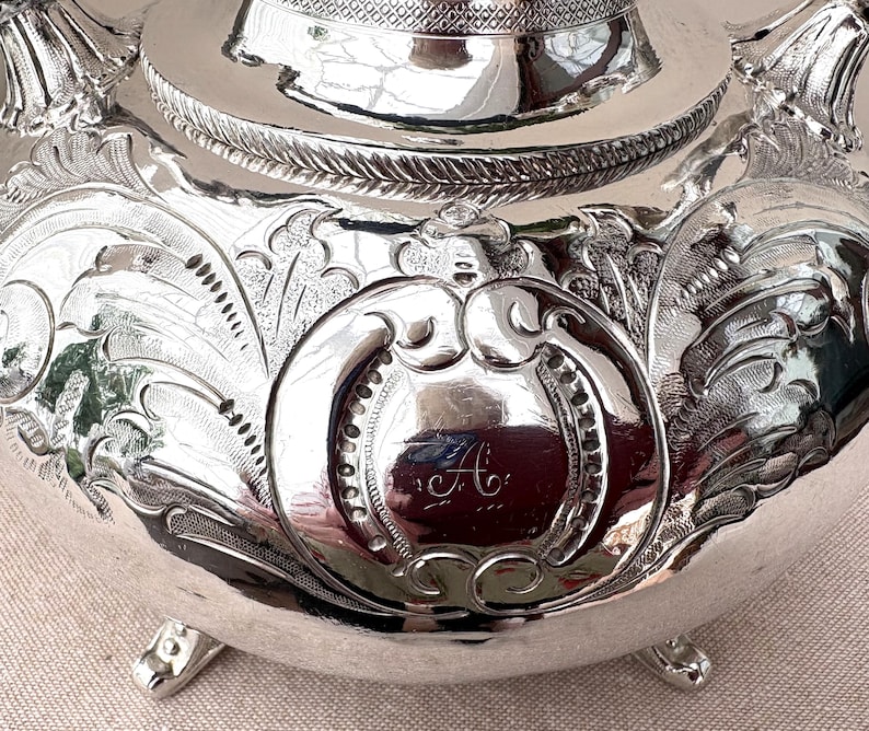 May include: A polished silver teapot featuring intricate embossed floral and scrollwork patterns across its rounded body. The center displays a circular cartouche engraved with the cursive letter A. It stands on small, ornate feet with a rope-like border.