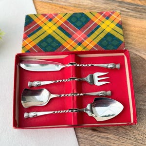 以下が含まれることがあります： 赤色の箱に入った、4つの銀色のサービング用具のセット。バターナイフ、フォーク、スプーン、ケーキサーバーが含まれています。各器具は、ねじれたハンドルと装飾的な端を持っています。蓋はチェック柄です。