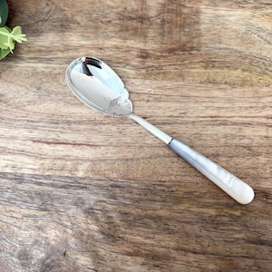 Op de afbeelding: Een zilveren serveerlepel met een ovale kom en een decoratief handvat. De kom van de lepel heeft een sierlijk ontwerp en de steel gaat over van zilver naar een parelmoerwit. De lepel ligt op een houten achtergrond.