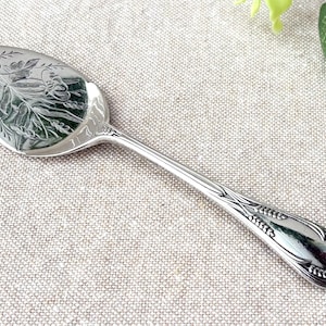 May include: A silver-toned metal serving spoon featuring an oval bowl with an engraved floral and wheat pattern. The handle is slender and tapers toward the end, decorated with a matching wheat motif along the edges and a central embossed design.