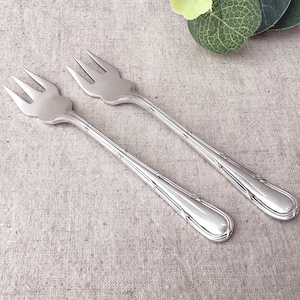 Peut inclure: Deux fourchettes à cocktail en métal argenté, chacune dotée de trois dents. Les manches présentent un motif gaufré décoratif avec des lignes verticales et une extrémité arrondie lisse. Les fourchettes ont une finition polie et un design fin.
