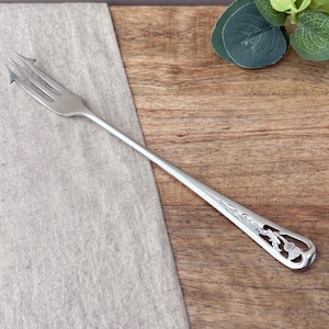 May include: A silver-toned metal fork featuring a long, slender handle with an openwork design at the end. The handle is decorated with an embossed thistle motif and intricate scrollwork patterns, while the fork head has three distinct tines.