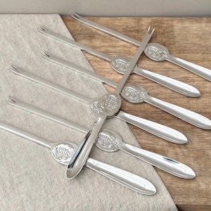 May include: A set of six silver-toned metal cocktail forks, each featuring a two-pronged tip and an oval handle embossed with a detailed pineapple design. The forks are arranged in a fan-like pattern, showcasing their polished metallic finish.