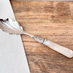 Puede incluir: Una cuchara de servir plateada con un cuenco en forma de concha y un mango con un agarre de nácar. La cuchara está sobre una superficie de madera junto a un paño blanco.