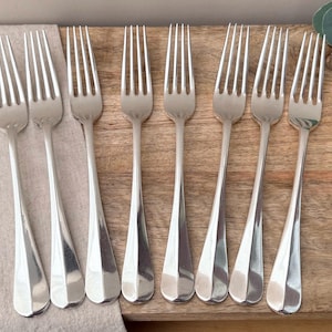 May include: A set of eight polished silver-toned metal forks arranged in a row. Each fork features a slender handle with a subtle faceted design, four long tines, and a smooth, reflective finish. The forks are uniform in size and shape.