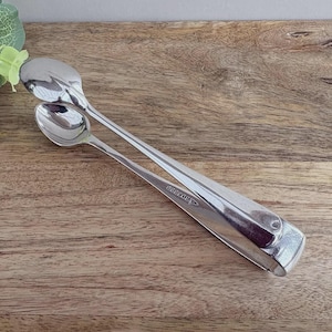 May include: Silver-colored sugar tongs with two spoon-shaped ends. The tongs are resting on a wooden surface. The tongs are designed to pick up sugar cubes or other small items. The tongs are a vintage or antique item.