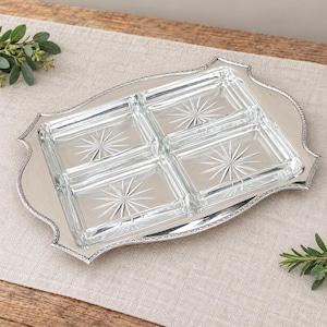 May include: A rectangular silver-toned serving tray with a decorative beaded edge, featuring four clear glass rectangular inserts. Each glass insert is etched with a central starburst pattern, providing a clear and segmented surface.