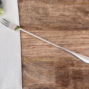Op de afbeelding: Een zilveren vork met een lange steel, rustend op een houten oppervlak naast een witte doek en groen. De vork heeft drie tanden en een gepolijste afwerking, ideaal voor het serveren van voorgerechten of desserts.