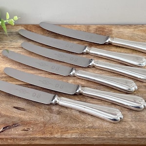 Könnte beinhalten: Sechs silberne Buttermesser mit verzierten Griffen liegen auf einer Holzoberfläche. Die Messer haben ein klassisches Design mit glänzendem Finish. Die Klingen sind glatt und die Griffe haben ein geriffeltes Muster. Die Messer sind ca. 20 cm lang.