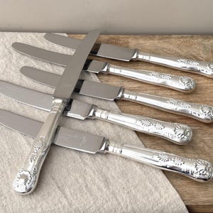 Könnte beinhalten: Ein Set aus fünf silberfarbenen Tafelmessern mit polierten Edelstahlklingen und verzierten, geprägten Griffen mit einem muschelartigen Motiv am Ende. Die Messer sind fächerförmig angeordnet, wobei ein Messer quer über den anderen liegt.