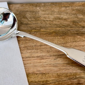 May include: A silver ladle with a round bowl and long handle, resting on a wooden surface. The handle has an engraved initial. The ladle is next to a white cloth.