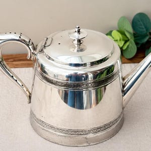 Op de afbeelding: Een gepolijste zilveren theepot met een scharnierend deksel met een centrale knop, een gebogen handvat met decoratieve perforaties en een taps toelopende tuit. Het lichaam is versierd met twee horizontale banden van reliëfpatronen.